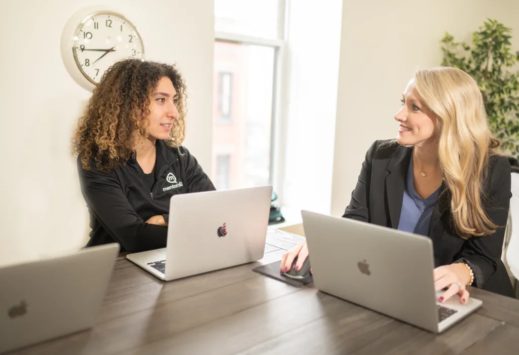 Two women working on laptops across from each other at an office table, engaged in a conversation. One woman has curly hair and wears a black shirt, while the other, with blonde hair, wears a business suit.