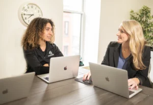 Two women working on laptops across from each other at an office table, engaged in a conversation. One woman has curly hair and wears a black shirt, while the other, with blonde hair, wears a business suit.