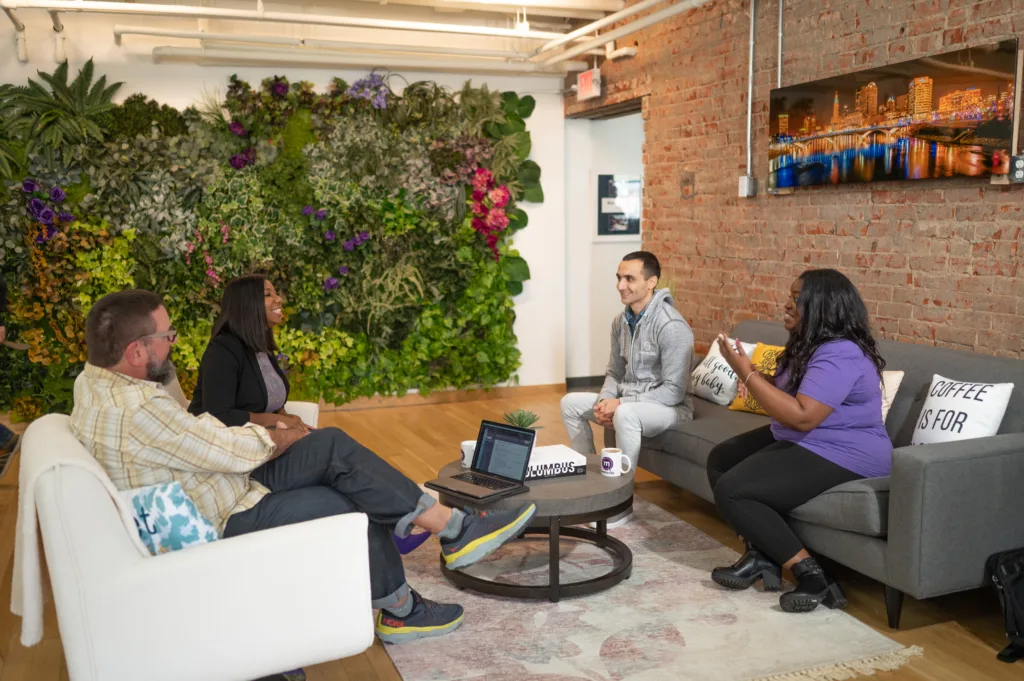 Photo of four professionals having a casual meeting in a modern office space. They are seated in a relaxed setting with a large living plant wall, a brick wall, and urban decor. They appear engaged in a lively conversation.